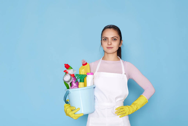 calm caucasian woman in gloves holding bucket with cleaning supplies isolated on blue background. - Zdjęcie, obraz