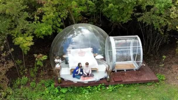 Прозрачный шатер пузыря при глампинге, пышный лес вокруг и внутри. Пара, сидящая под одеялом, держа чашки и разговаривая - Кадры, видео
