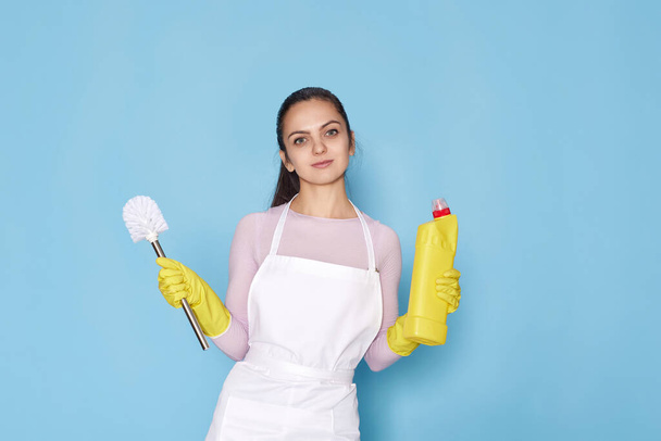 woman in yellow rubber gloves and cleaner apron holding toilet brush and bottle of detergent on blue background. - Photo, image