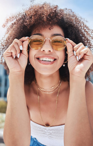 Portrait of young trendy beautiful mixed race woman with an afro smiling and posing alone outside. Hispanic woman wearing sunglasses and feeling happy. Fashionable African American woman in the city. - Foto, Imagem