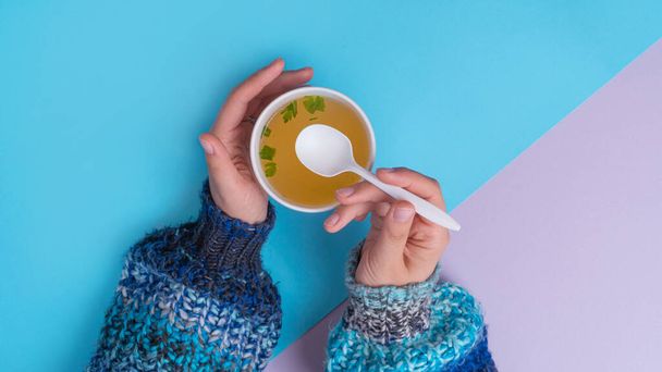 Famele in sweater eating a Chicken broth soup in takeaway cup with spoon. Minimalistic Blue and purple background - Photo, Image