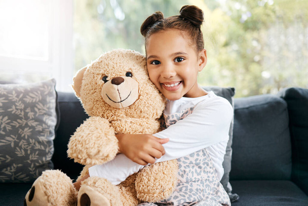 Porträt eines entzückenden kleinen hispanischen Mädchens, das zu Hause einen Teddybär hält. Niedliche gemischte Rassenmädchen spielen mit ihrem Spielzeug, während sie auf der Couch in der Lounge sitzen. - Foto, Bild
