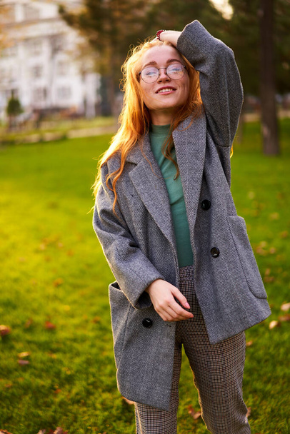 Redhead smiling pretty woman walking in park on sunny autumn day. Elegant female poses in stylish authentic outfit has fun laughing on green lawn with vivid foliage, leaves. Girl enjoys a fall season. - Фото, изображение