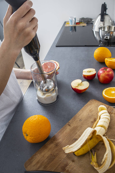 Mujer usando una licuadora de manos para mezclar frutas frescas en la cocina. - Foto, Imagen