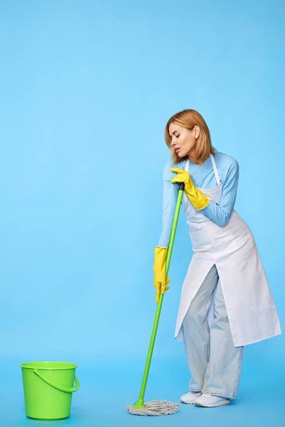 mujer alegre feliz en guantes de goma y delantal más limpio lavando piso con fregona sobre fondo azul. Longitud completa - Foto, imagen