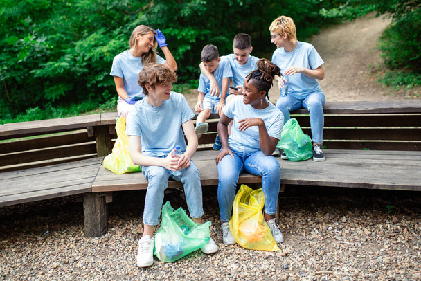Spontanes Bild einer kleinen Gruppe Freiwilliger mit Handschuhen und Müllsäcken, die im gerade geputzten Park Halt machen. Alle tragen blaue T-Shirts und sind stolz und glücklich auf das, was sie getan haben - Foto, Bild