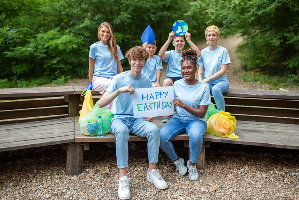 幸せなボランティアは、「幸せな地球の日」のメッセージでプラカードを保持します。ボランティア、慈善、清掃、人々と生態学の概念-公園内のゴミ袋の清掃エリアを持つ幸せなボランティアのグループ - 写真・画像