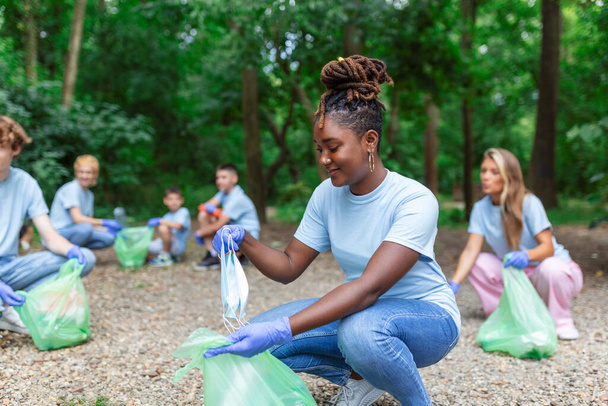 Een groep mensen maakt samen schoon in een openbaar park, om het milieu te beschermen. Vrouw op de voorgrond met een vuilniszak in haar hand reinigt het park - Foto, afbeelding