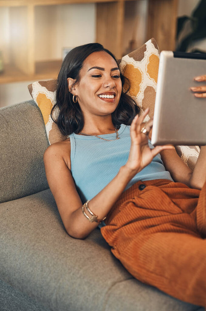 Smiling mixed race woman browsing internet on digital tablet at home. Happy hispanic lying down on living room sofa alone and using technology. Relaxed woman scrolling and searching online on weekend. - Foto, Bild