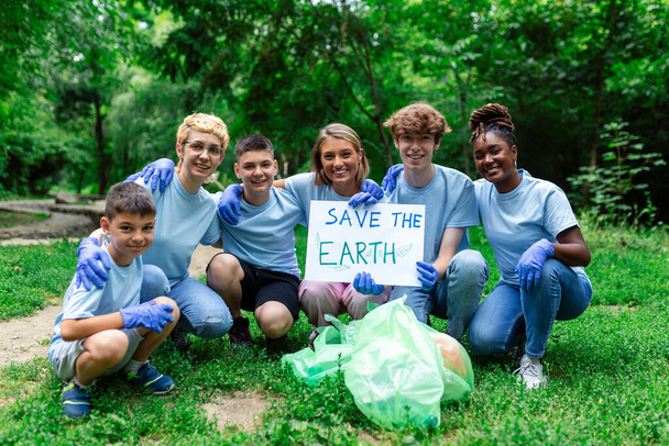 Διαφορετική ομάδα ανθρώπων μαζεύει σκουπίδια στην εθελοντική κοινωνική υπηρεσία του πάρκου. Ευτυχείς διεθνείς εθελοντές που κρατούν πλακάτ με το μήνυμα "Σώστε τη Γη". - Φωτογραφία, εικόνα