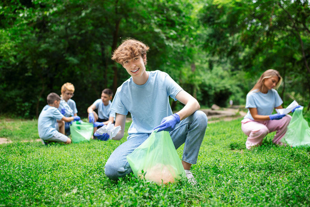 Ευτυχισμένοι άνθρωποι της κοινωνικής υπηρεσίας να καθαρίζουν το τοπικό πάρκο. Έχουν μια σακούλα σκουπιδιών και γεμίζουν με σκουπίδια που έχουν βρει στο πάρκο. Είναι δύο γονατιστοί και άλλοι πίσω τους - Φωτογραφία, εικόνα
