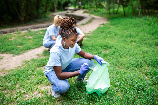 Dobrovolnictví, charita a koncepce čistého životního prostředí. Šťastná africká žena a skupina dobrovolníků s čistírnou odpadkových pytlů v parku, kopírovací prostor - Fotografie, Obrázek