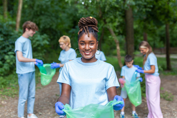 Portrait d'une belle femme avec un petit groupe de bénévoles en arrière-plan avec des gants et des sacs à ordures qui nettoient le parc municipal - concept de préservation de l'environnement et d'écologie. Tous portant un t-shirt bleu - Photo, image