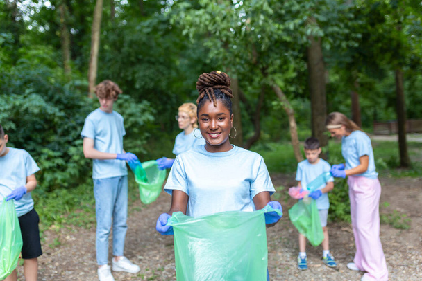 The young adult woman, who is one of a diverse group of volunteers, takes time to smile for the camera. Hold garbage bag and looking at camera - Photo, Image