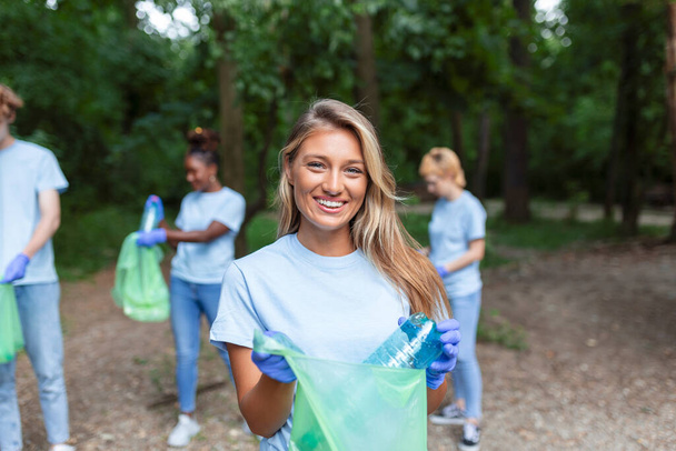 Die junge erwachsene Frau, die zu einer vielfältigen Gruppe von Freiwilligen gehört, nimmt sich Zeit, um für die Kamera zu lächeln. Müllsack halten und in die Kamera schauen - Foto, Bild