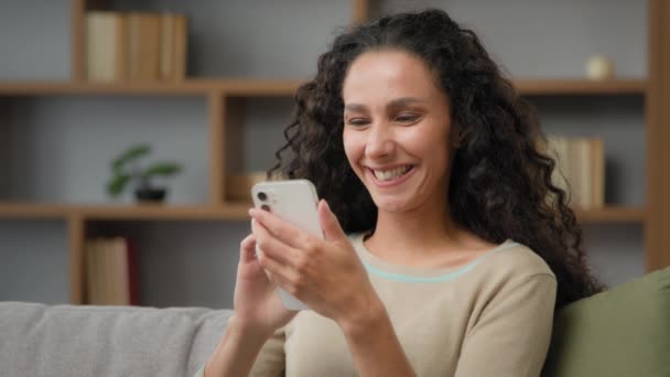 Happy smiling toothy laughing female Caucasian Latino Hispanic woman with curly hair laugh with funny video in smartphone holding phone cellular watching mobile fun humorous app online at home couch - Footage, Video