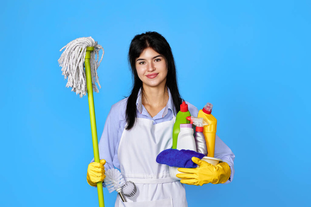 pretty woman in yellow rubber gloves and cleaner apron holding bucket of detergents and mop on blue background. - Foto, imagen