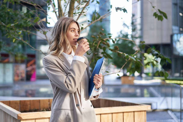 Schöne lächelnde Geschäftsfrau trinkt in ihrer Arbeitspause Kaffee und steht auf der Straße. - Foto, Bild