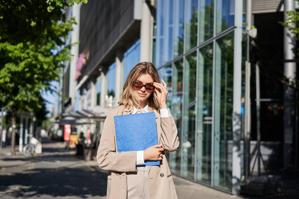 Портрет корпоративной женщины в солнечных очках и бежевом костюме, держащей синюю папку с офисными документами, идущей на работу, гуляющей по улице в солнечный день. - Фото, изображение