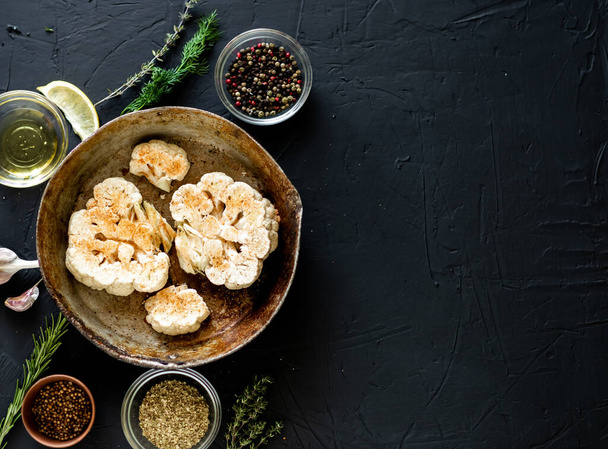 Cauliflower steak cooking. Raw cauliflower sprinkled with spices lies in a frying pan. Olive oil, herbs, various spices nearby. Dark background. Copyspace. - Foto, Imagen