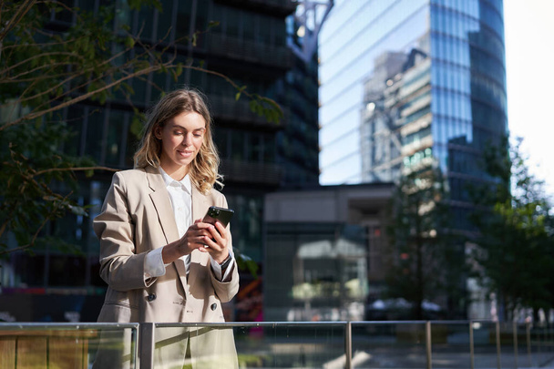 Молодая деловая женщина, стоящая на улице. Офис работника с помощью мобильного телефона, глядя на смартфон. - Фото, изображение