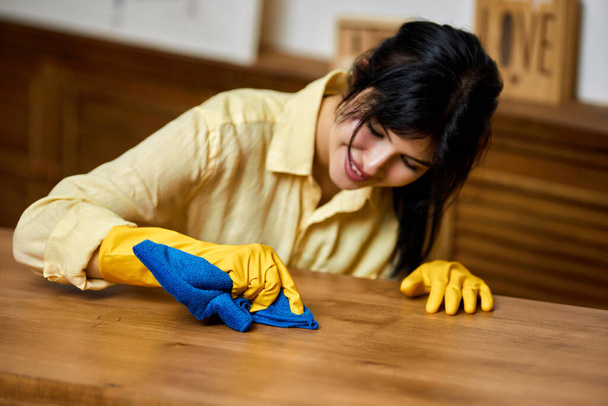brunette woman in yellow gloves using a rag while cleaning furniture at home - Fotó, kép