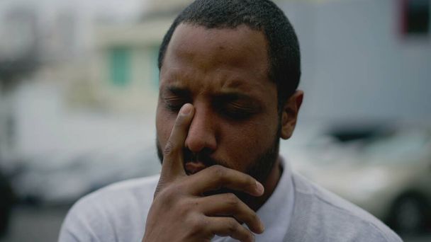 Niespokojny młody człowiek odczuwający rozpacz i negatywne myśli. Afroamerykanin cierpiący na chorobę psychiczną, stojący na ulicy i zakrywający twarz ręką - Zdjęcie, obraz