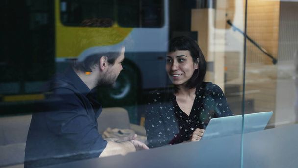 Zwei junge Leute unterhalten sich über die Arbeit vor Laptop und Arbeitsplatz, betrachtet durch die Spiegelung der Fenster. Geschäftsfrau und Mann diskutieren im Gespräch - Foto, Bild