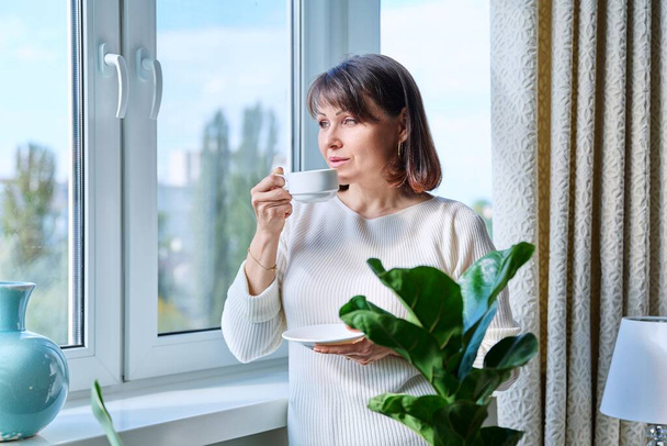 Middle aged woman at home near window with cup of coffee. Smiling calm mature woman looking out the window, morning coffee, lifestyle, 40s people concept - Φωτογραφία, εικόνα