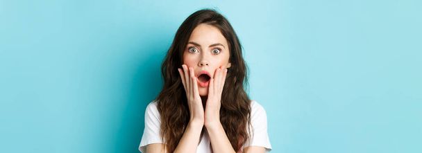 Oh my gosh. Close up portrait of shocked girl face, touching head and staring at camera startled, hear bad news, standing over blue background. - Fotografie, Obrázek