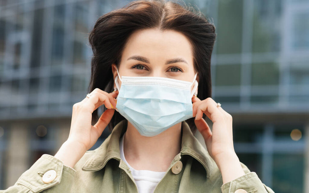 Young Woman Putting a Protective Face Mask on Her Face Because of Pandemic, Portrait of Brunette Girl Using Earphones Standing Outdoors Against City Buildings - Photo, Image