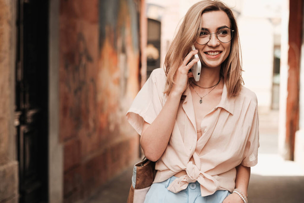 Χαρούμενη κομψή γυναίκα σε γυαλιά Χρησιμοποιώντας Smartphone και χαμογελώντας, κομψό ξανθό κορίτσι με γυαλιά ηλίου στέκεται έξω στο δρόμο έχοντας συνομιλία μέσω του κινητού τηλεφώνου - Φωτογραφία, εικόνα