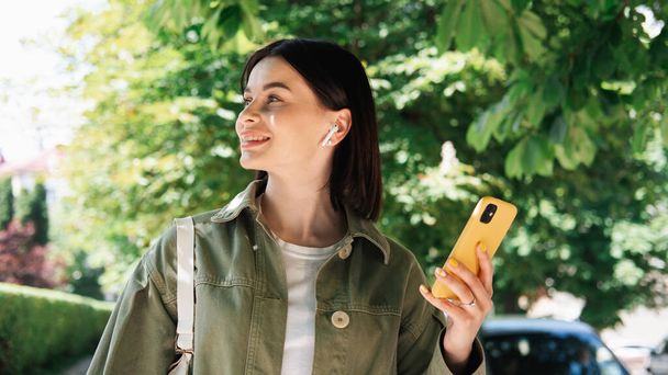 Νεαρή γυναίκα κρατώντας Smartphone στο χέρι, ενώ το περπάτημα στην πόλη, Brunette κορίτσι χρησιμοποιώντας το κινητό τηλέφωνο και ακουστικά κοιτάζοντας μακριά και χαμογελώντας - Φωτογραφία, εικόνα