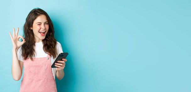 Online shopping. Cheerful cute girl winking at you, smiling and showing okay sign after using smartphone app, recommending internet shop or social media page, blue background. - Photo, Image