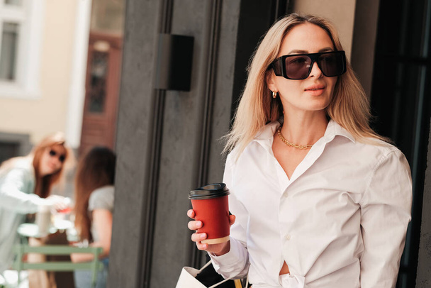 Elegancka kobieta wychodzi z kawiarni, trzymając filiżankę kawy w dłoni i patrząc w dal, pewna siebie przedsiębiorczyni nosząca okulary przeciwsłoneczne, koncepcja kawy to Go - Zdjęcie, obraz