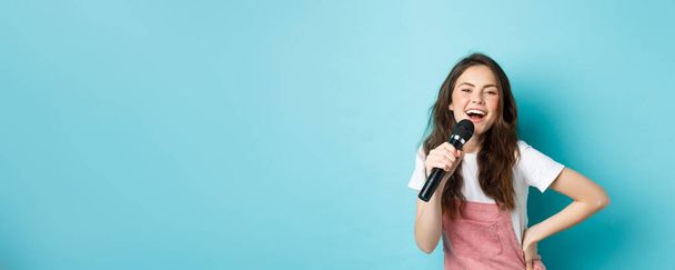 Веселая молодая женщина поет караоке, держит микрофон и улыбается, веселится, стоя на синем фоне. - Фото, изображение