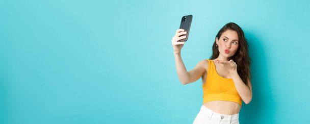 Technologie en lifestyle concept. Stijlvolle mooie vrouw poseren voor selfie, het verzenden van luchtkus op camera, met behulp van foto filters app op mobiele telefoon, staande tegen een blauwe achtergrond. - Foto, afbeelding