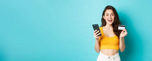 Young happy woman making online order, shopping with credit card and smartphone, look amazed at camera, standing over blue background. - Photo, Image