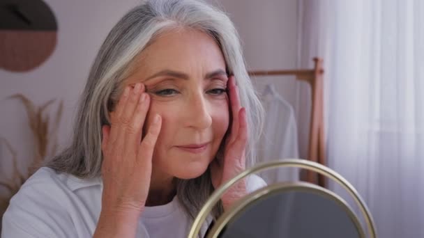 Senior gray-haired older 60s woman 50s lady granny with wrinkled beautiful aging face with perfect smooth skin looking at mirror reflection touching cheeks massaging skincare cosmetology treatment - Footage, Video