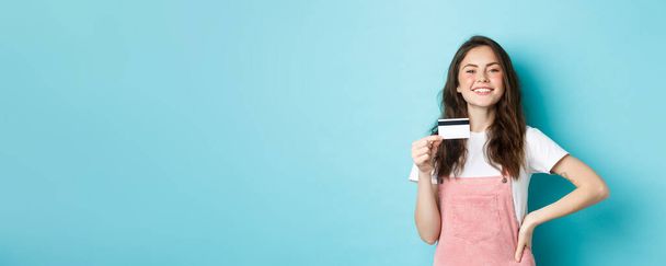 Trots lachend meisje kijkt opschepperig, toont haar plastic creditcard, gaat om iets te kopen, winkelen contactloos, staande over blauwe achtergrond. - Foto, afbeelding