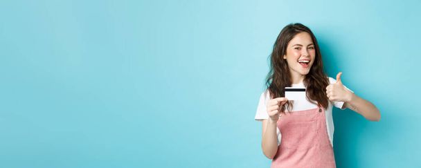 Zufriedene schöne moderne Mädchen zeigt Plastikkreditkarte und Daumen nach oben, empfehlen Bank oder Geschäft, steht über blauem Hintergrund. - Foto, Bild