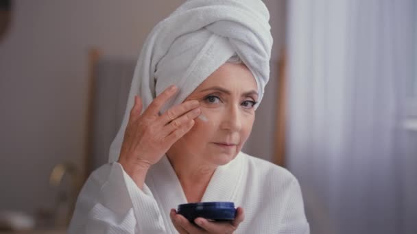 Caucasian old 60s woman 50s lady grandmother aged model skin care in bathroom wears bathrobe and towel on head applies moisturizing cream with collagen vitamins anti-wrinkle product to cheeks face - Footage, Video