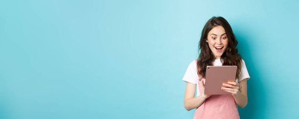 Image of excited young woman stare surprised at digital tablet screen, found something cool online, standing against blue background. - Фото, зображення