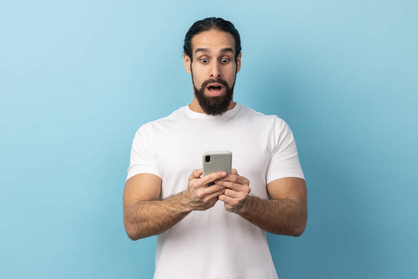 Portrét překvapeného muže s vousy v bílém tričku, který vyjadřuje šok při používání mobilního telefonu, hledá na webu, ohromující zprávy. Vnitřní studio záběr izolované na modrém pozadí. - Fotografie, Obrázek