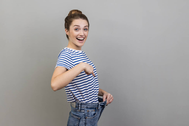 Attractive woman wearing striped T-shirt have willpower to loose weight, pointing at slim waist and pointing finger on big trousers, diet conception. Indoor studio shot isolated on gray background. - Фото, изображение