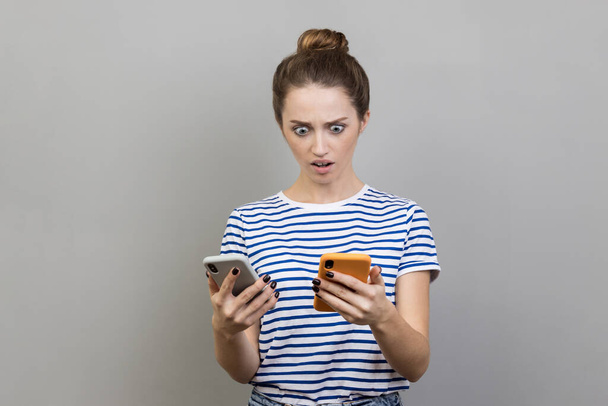 Retrato de una mujer sorprendida con una camiseta a rayas usando dos teléfonos inteligentes, mirando a la pantalla con ojos grandes, sorprendida. Estudio interior plano aislado sobre fondo gris. - Foto, imagen