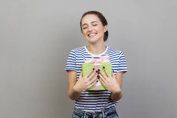 Portrait of happy smiling pleased woman wearing striped T-shirt embracing green wrapped present box, being satisfied of such a great gift. Indoor studio shot isolated on gray background. - Φωτογραφία, εικόνα