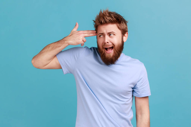 Depression, suicide gesture. Portrait of crazy bearded man standing with finger gun pointed to head, shooting himself with hand pistol. Indoor studio shot isolated on blue background. - Fotoğraf, Görsel
