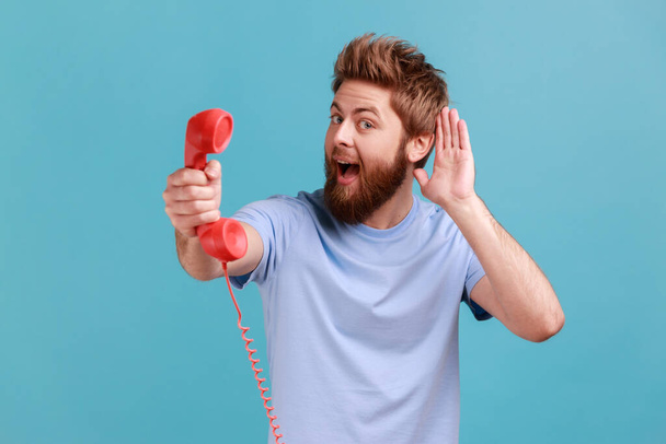 Portrait of bearded man holding and showing retro phone handset to camera, asking to answer phone, keeps hand near ear, hearing your talking. Indoor studio shot isolated on blue background. - Fotoğraf, Görsel