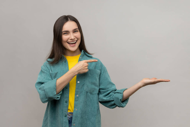 Portrait of happy woman presenting copy space on her palm, showing empty place for commercial text or goods, wearing casual style jacket. Indoor studio shot isolated on gray background. - Foto, immagini